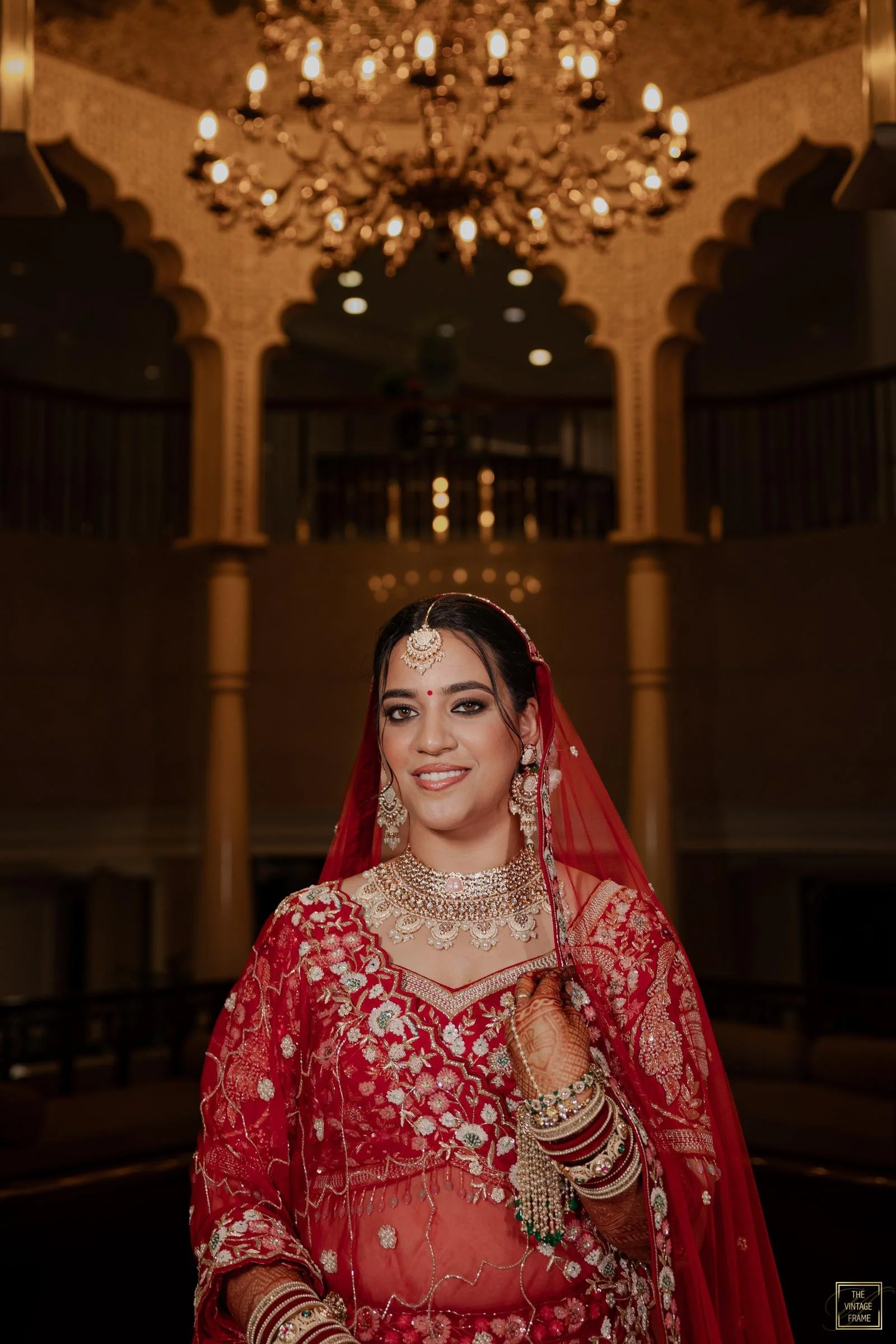 A smiling woman dressed in a traditional red bridal outfit with intricate embroidery and silver jewelry, standing in an ornate hall with a grand chandelier overhead.
