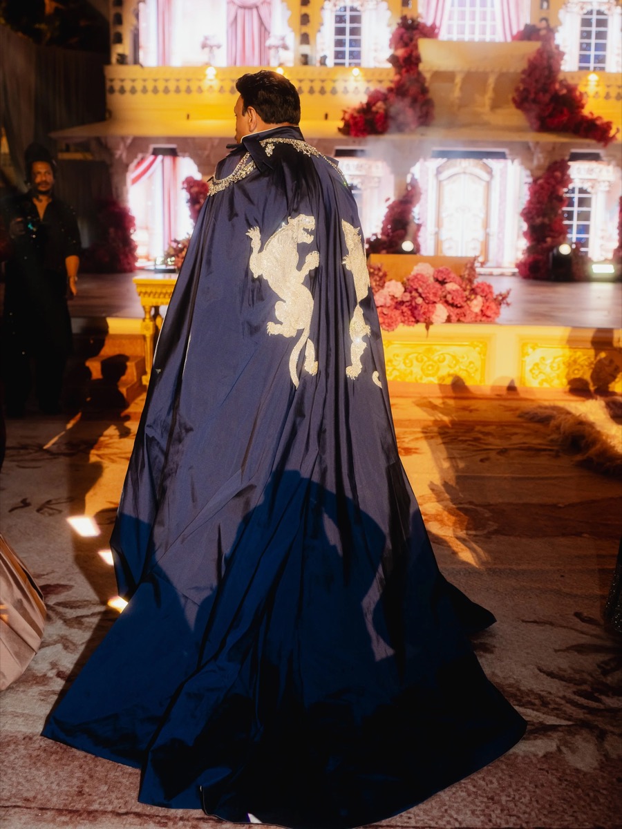 A man wearing a navy blue cape with golden lion designs, standing on a decorated stage with pink flowers and ornate architecture.