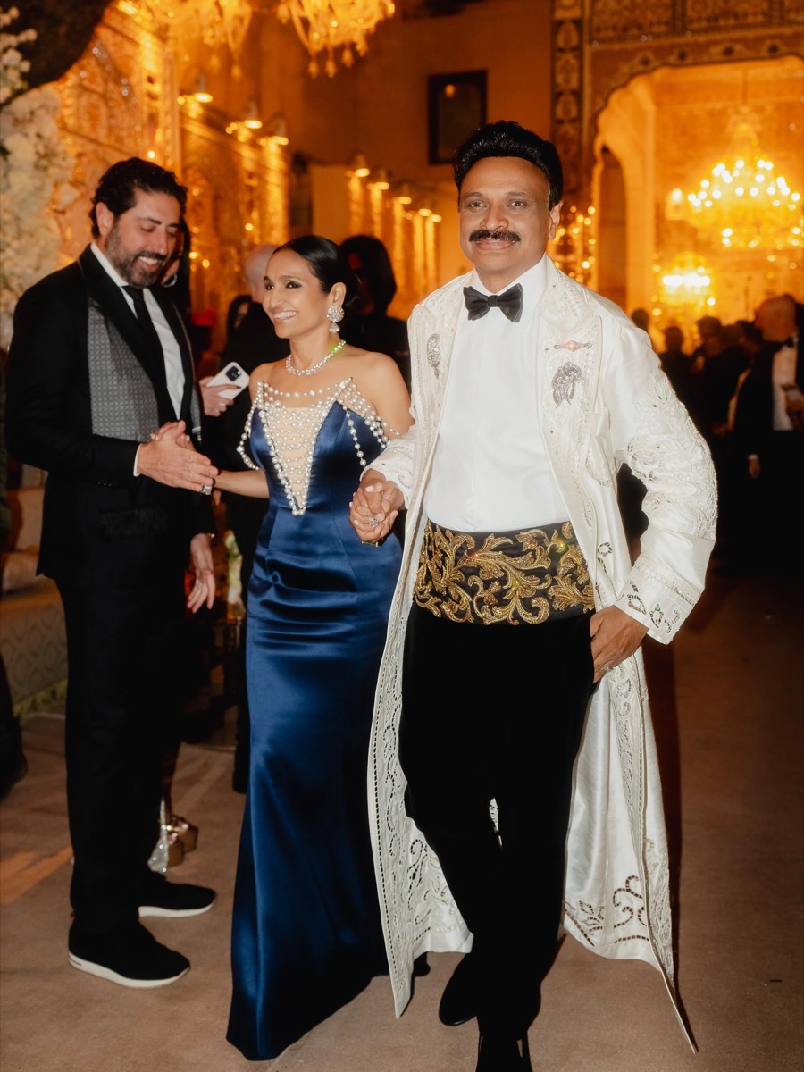 People at a formal event, dressed in elegant evening attire, with ornate decorations and warm lighting in the background.