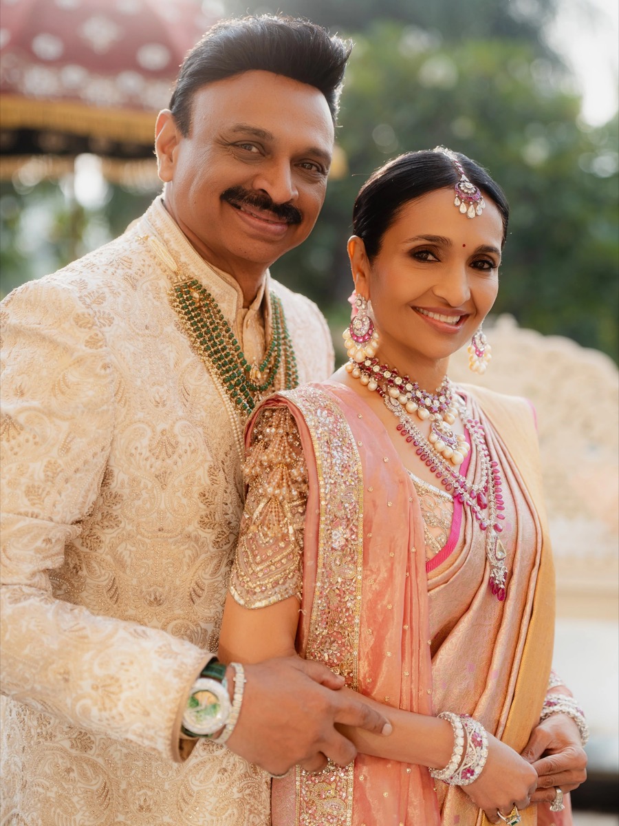 A smiling couple dressed in traditional Indian wedding attire outdoors, with the man wearing a cream-colored embroidered sherwani and jewelry, and the woman in a peach and pink saree with elaborate jewelry, including necklaces, earrings, and a headpi