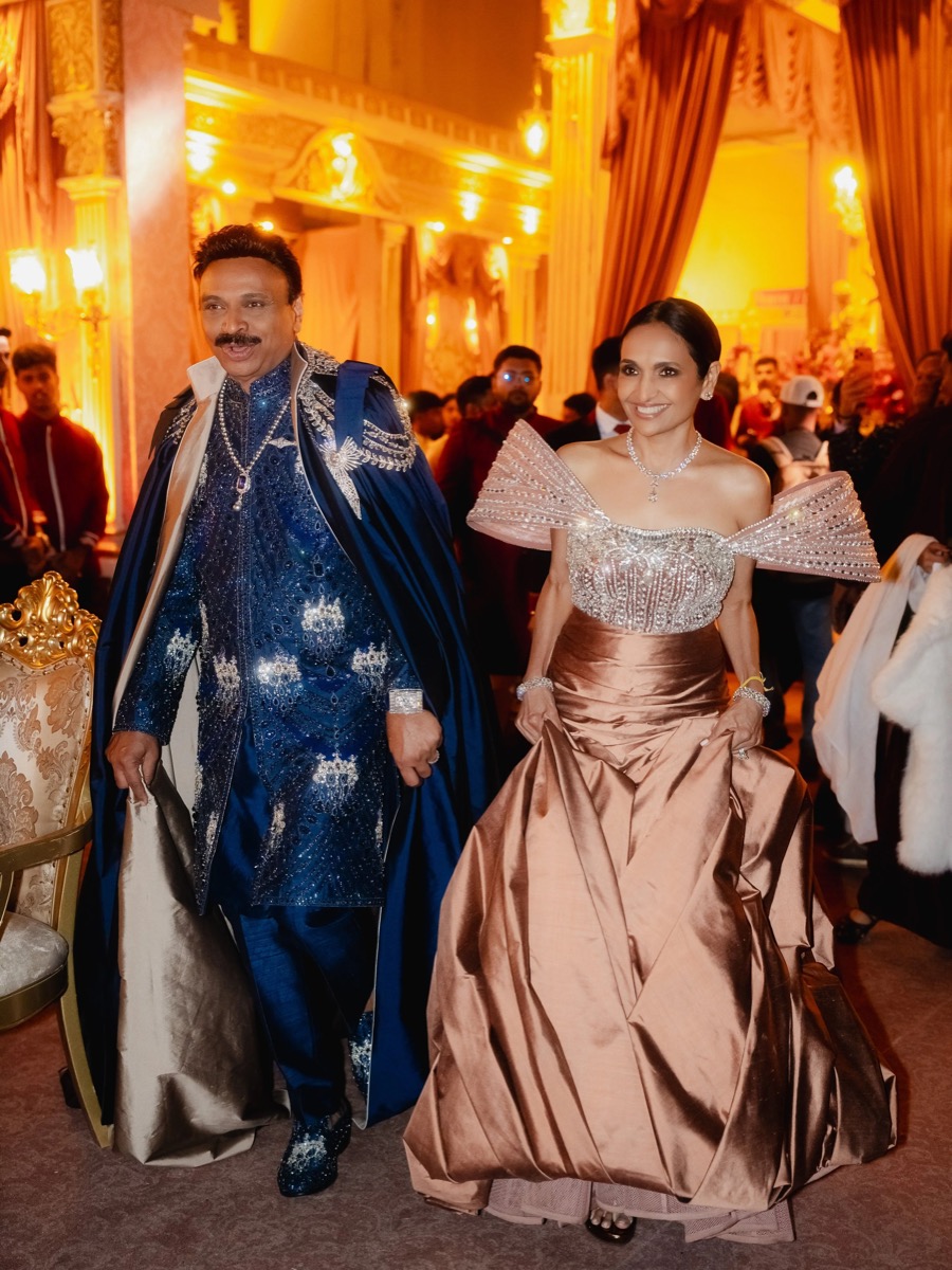 A man and woman dressed in elaborate, formal attire at a lavish event, with a grand, gold-lit interior and many people in the background.