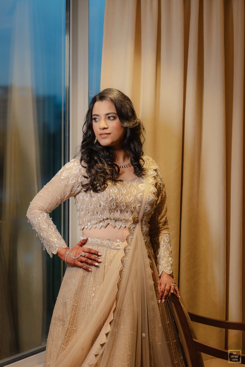 A woman dressed in a beige embroidered traditional outfit, standing beside a window with curtains, looking to her right with one hand on her waist and the other resting on a wooden railing.