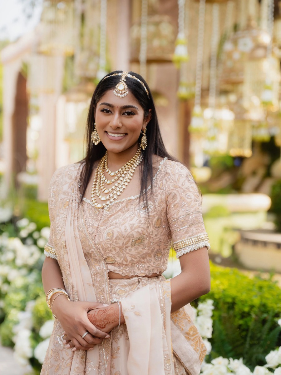 A woman dressed in traditional Indian bridal attire, smiling outdoors, wearing gold jewelry and intricate henna on her hands.
