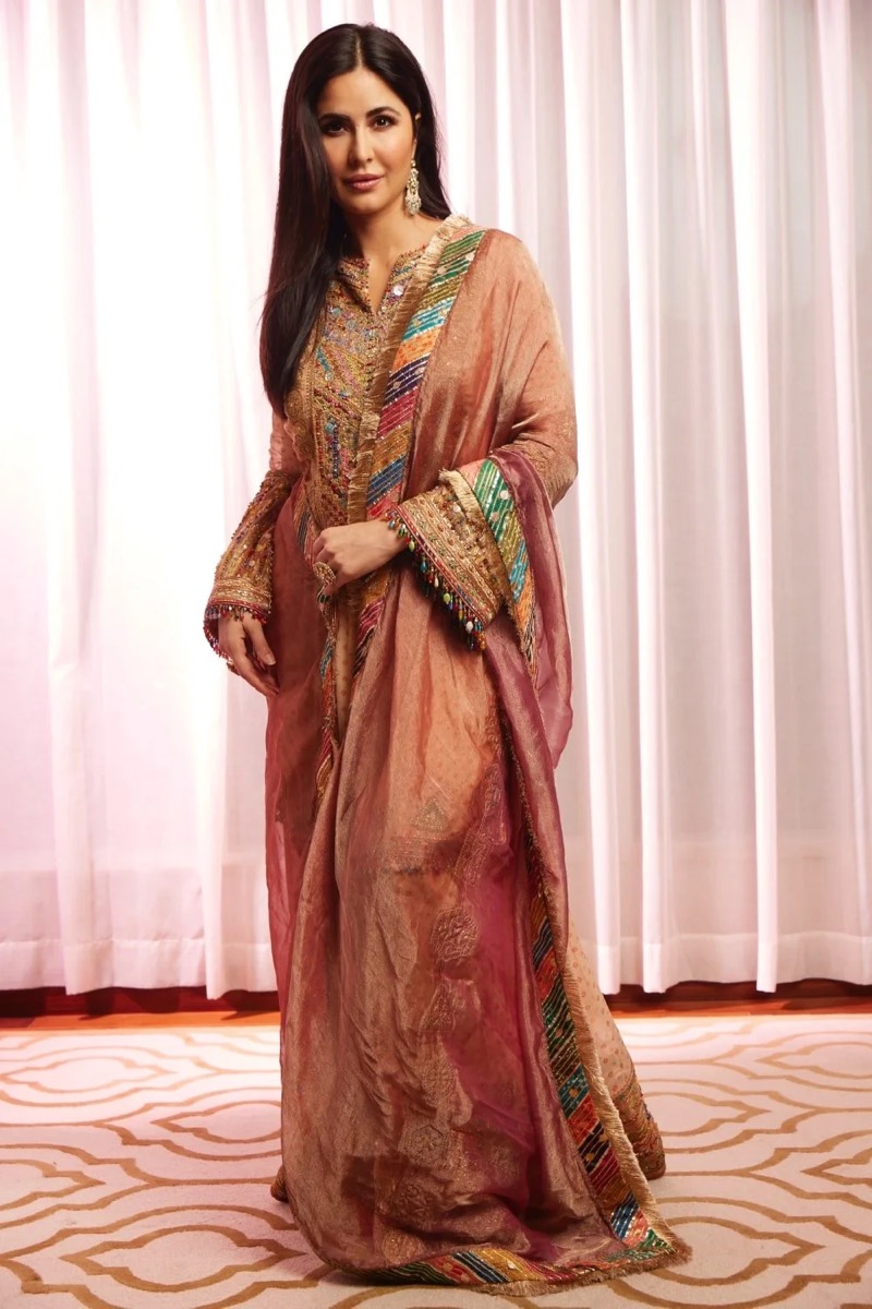 A woman wearing a traditional multicolored embroidered dress with a pink and gold draped shawl, standing on a patterned carpet in front of white curtains.