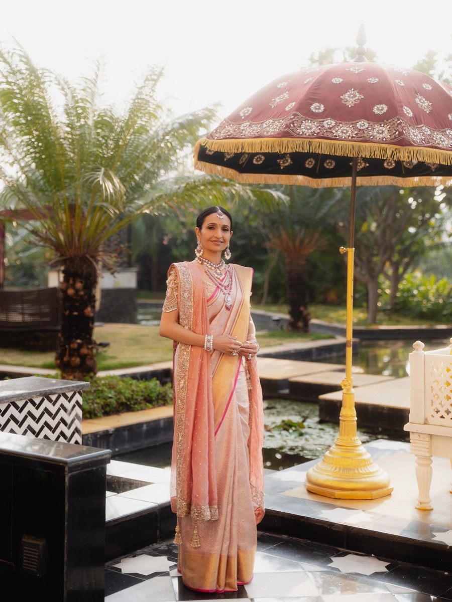 Woman dressed in traditional Indian attire standing outside near a decorative umbrella and water feature, surrounded by lush greenery.