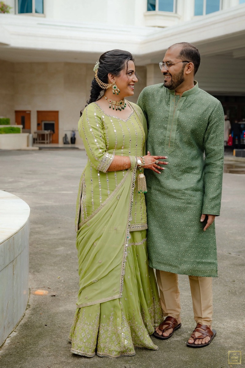 A woman and a man dressed in traditional Indian attire, standing close and smiling at each other outside a building.