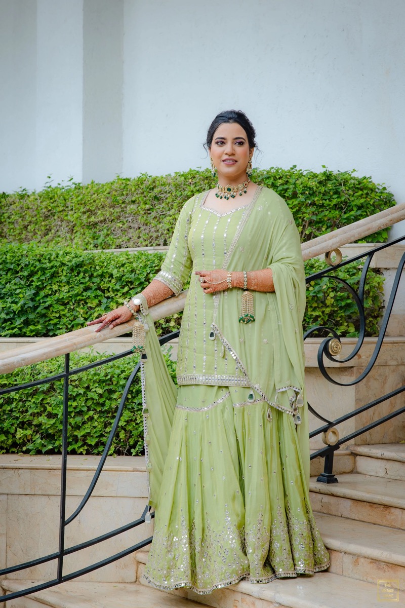 A woman dressed in a traditional lime green Indian outfit, adorned with jewelry, standing on a staircase outdoors with greenery and a white wall in the background.