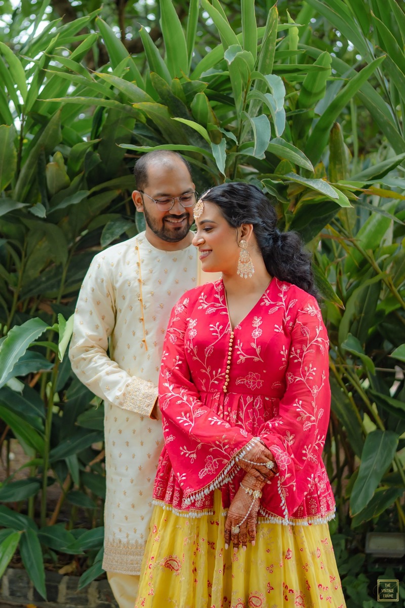 A couple in traditional Indian attire smiling and posing in front of green foliage.