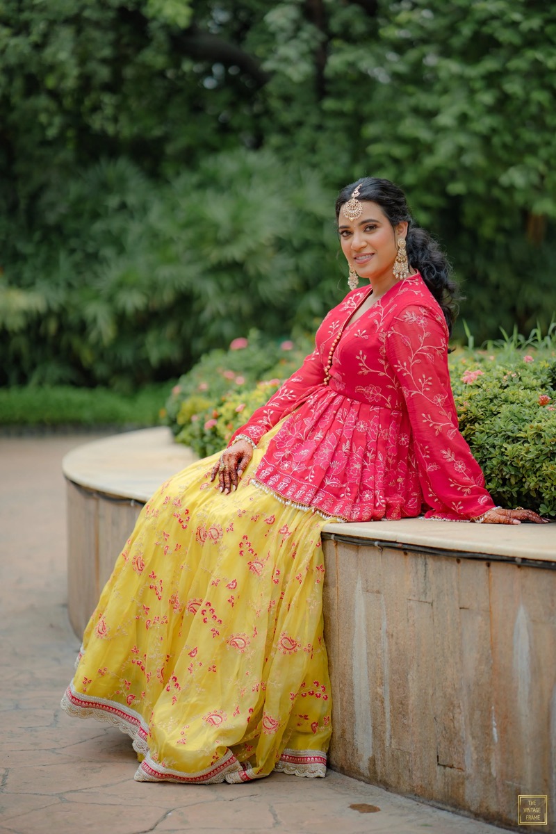 A woman dressed in traditional South Asian attire, sitting on a stone bench outdoors with greenery in the background, smiling at the camera.