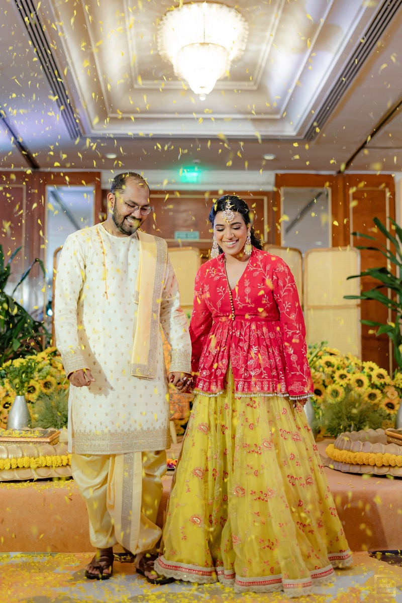 A bride and groom dressed in traditional Indian wedding attire, standing hand in hand amidst falling yellow flower petals, smiling happily during their wedding celebration.