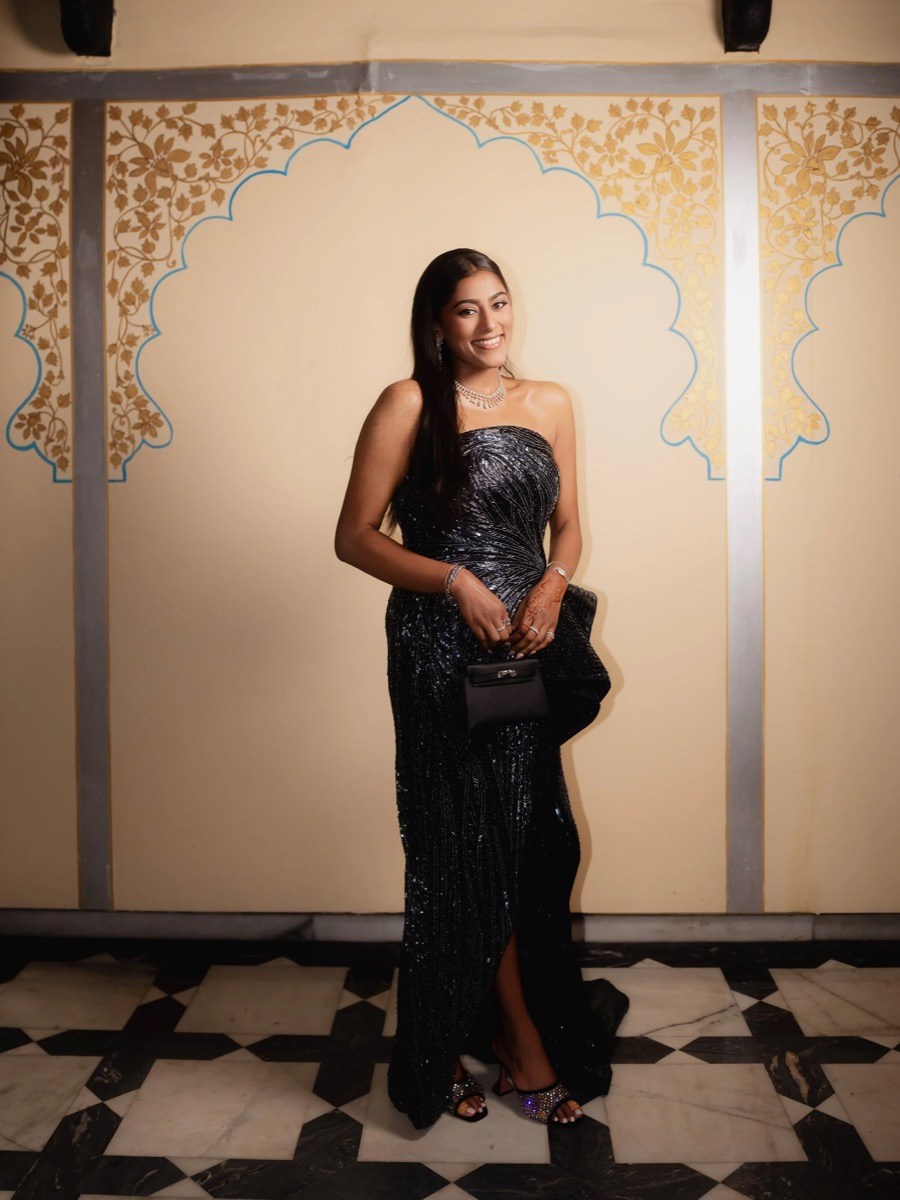 A woman in a black, sparkling evening gown holding a black clutch purse, standing in front of a decorated wall with gold floral patterns, smiling at the camera.