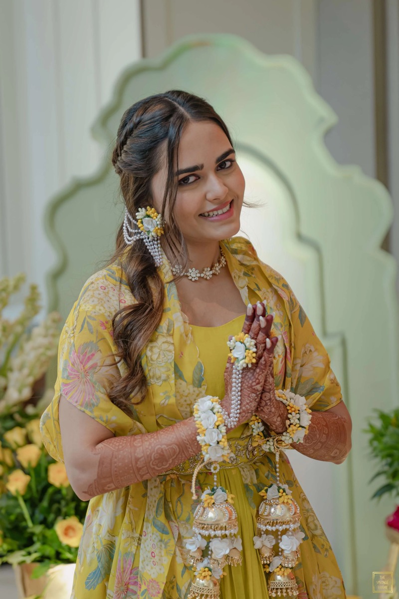 A woman dressed in yellow traditional attire, smiling, with henna on her hands, jewelry, and floral accessories, standing in front of a decorative background.
