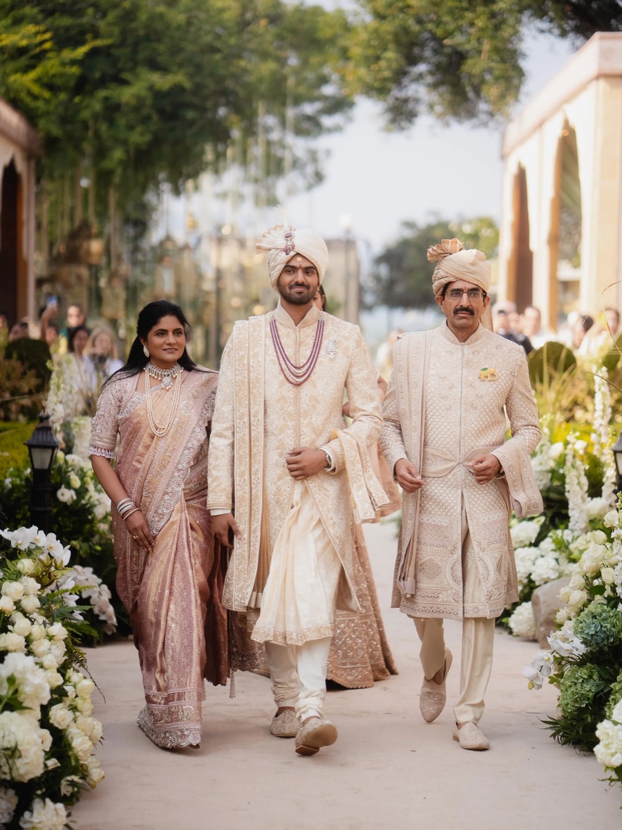 A group of people in traditional Indian wedding attire walking outdoors, including the bride and groom, and two men. They are surrounded by floral decorations and greenery.