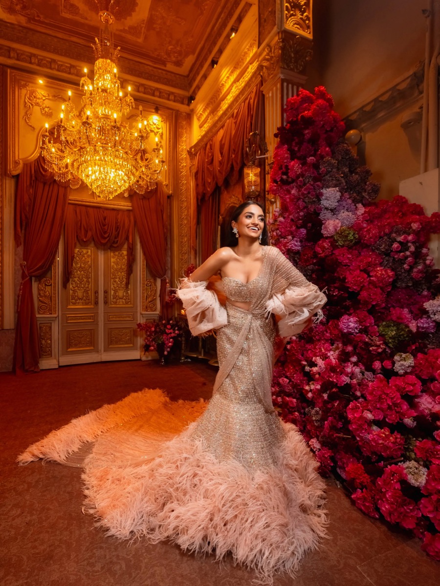 A woman in a glittering, peach-colored gown with fluffy feather details at the bottom, standing in a luxurious, ornately decorated room with a large chandelier and a wall of pink and purple flowers.