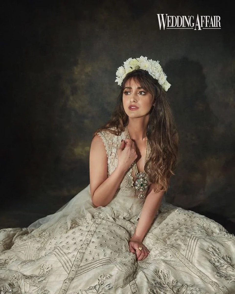 A woman in vintage wedding attire sitting on the floor with a dark, cloudy background, wearing a floral crown and jewelry, looking thoughtfully to the side.