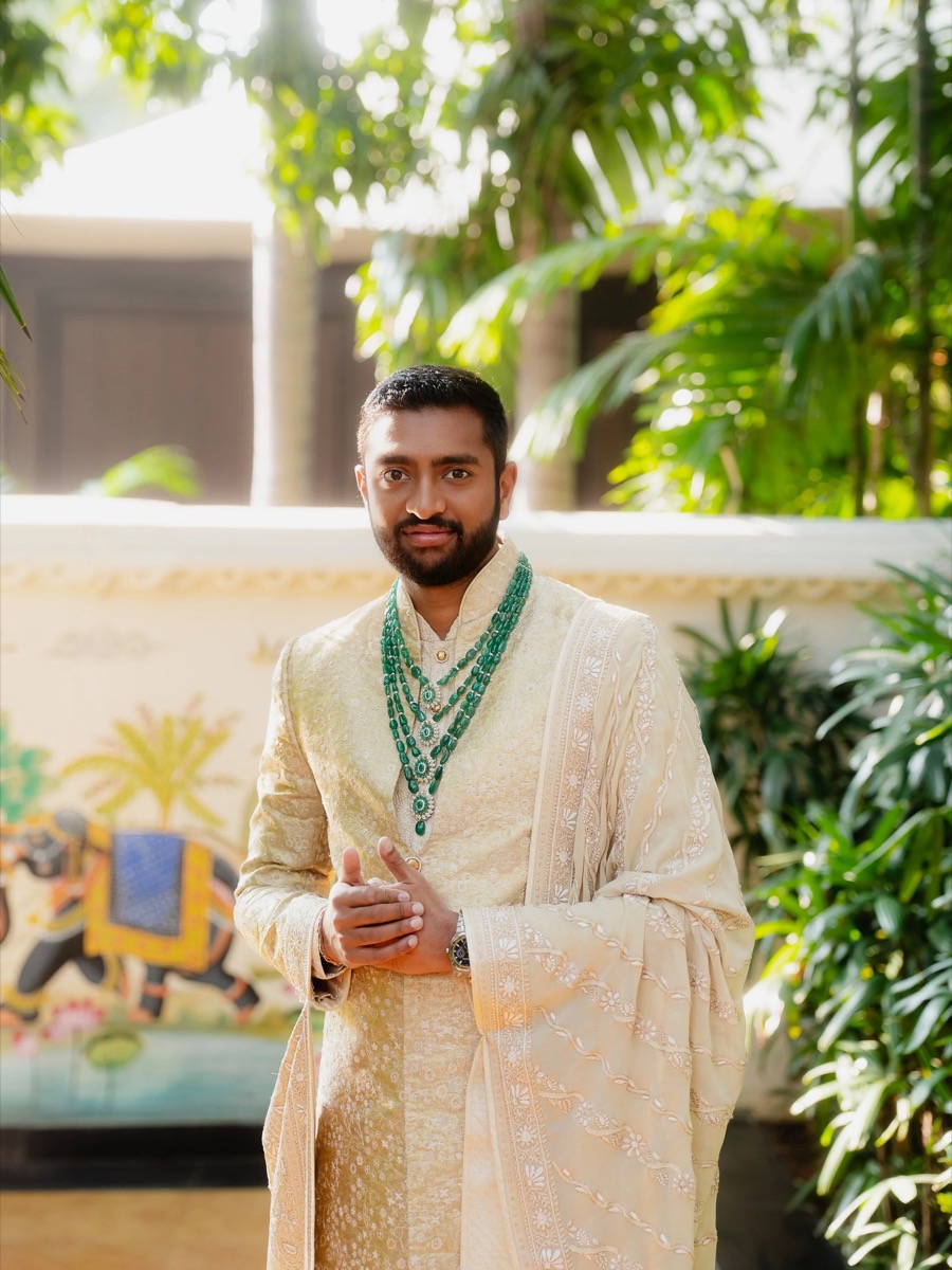 A man dressed in traditional Indian attire, wearing a cream-colored embroidered kurta with a matching dupatta, adorned with multiple green gemstone necklaces, stands outdoors in front of green foliage and a decorative backdrop.