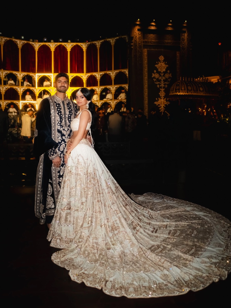 A couple dressed in traditional and elegant attire at a night-time event with ornate decor and a stage backdrop.