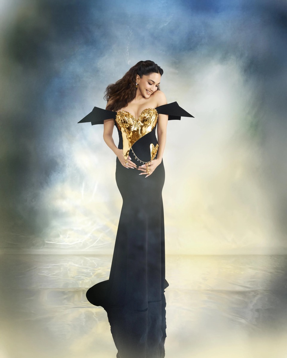 A woman wearing a black gown with gold decorative elements and black shoulder accents, smiling and looking down, standing on a reflective surface against a colorful, cloudy background.