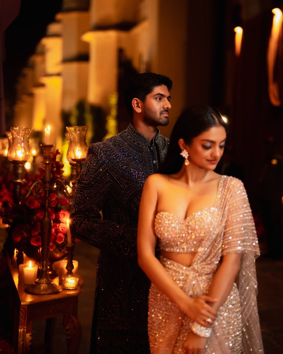 A man and woman dressed in elegant, traditional Indian attire at a festive event, with candles and flowers in the background.