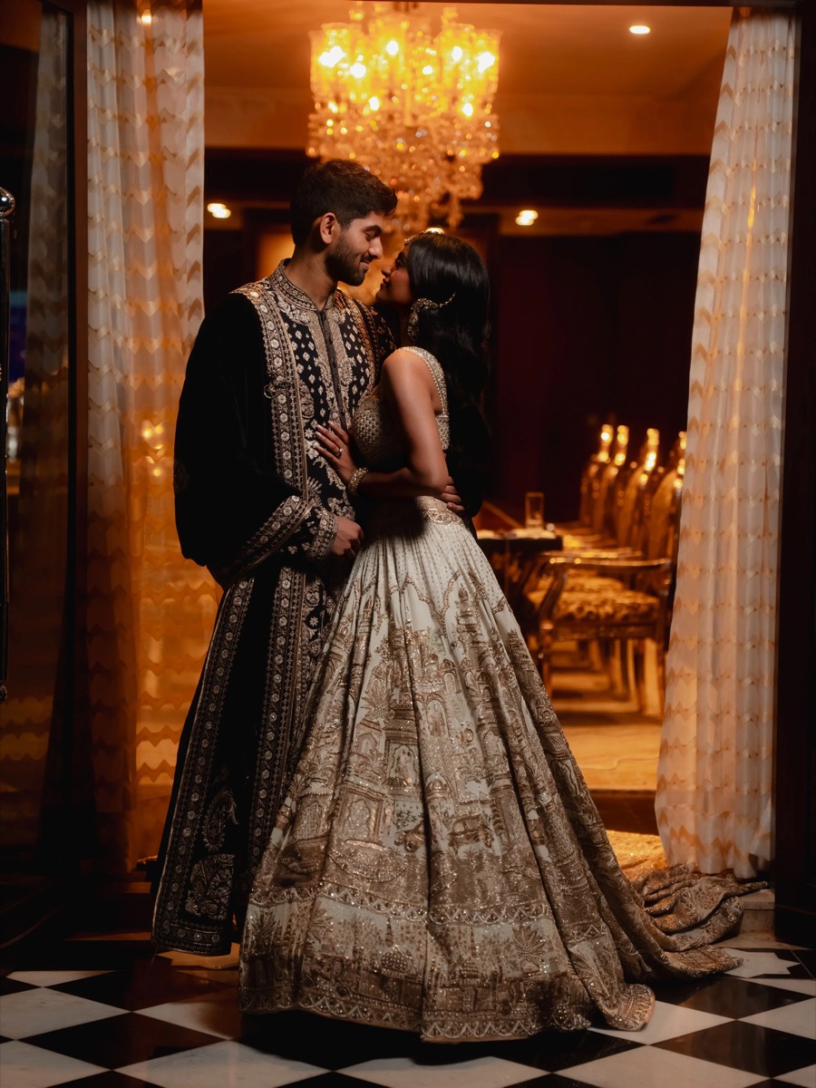 A couple dressed in traditional Indian wedding attire standing close together in a warmly lit room with a chandelier in the background.