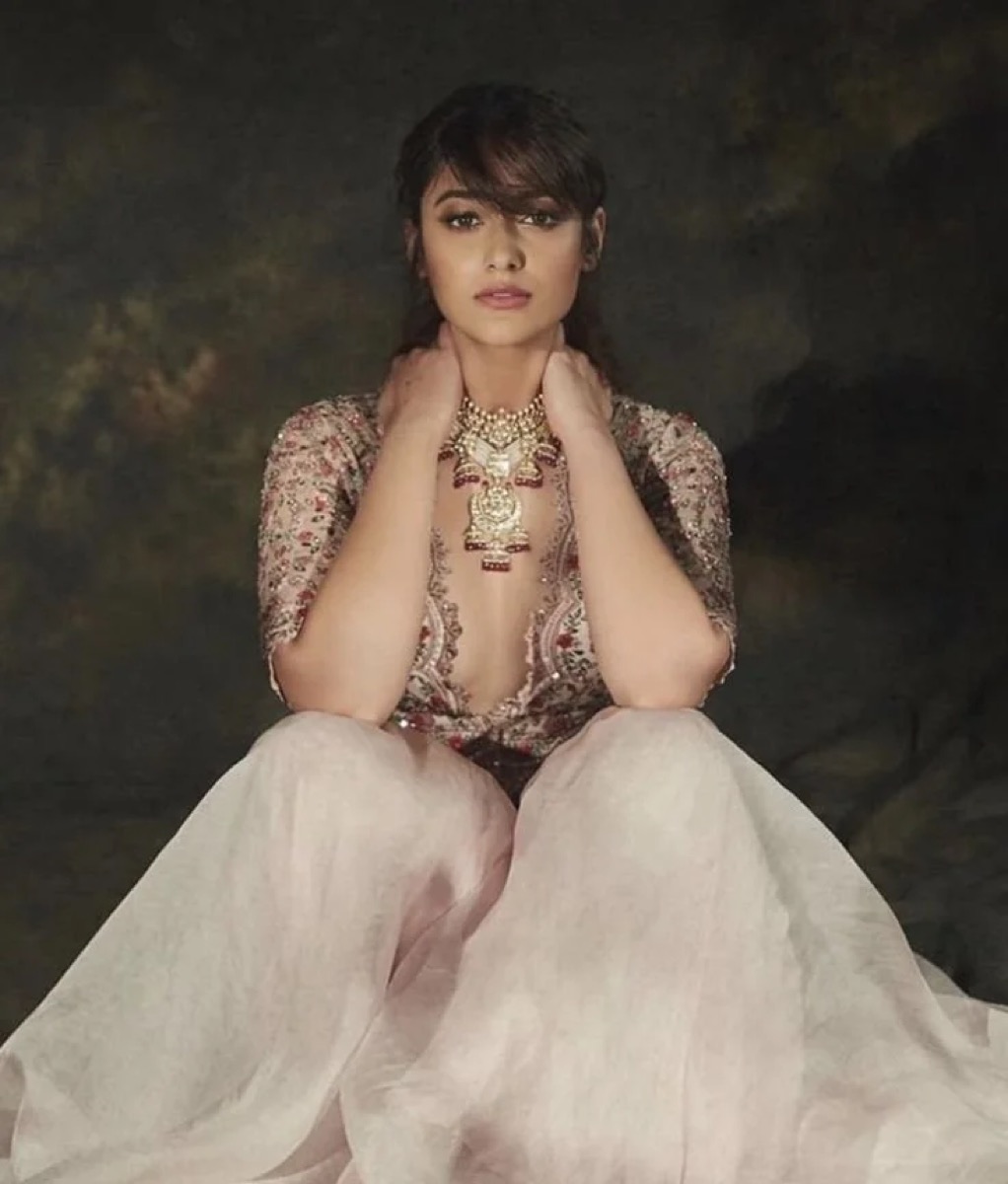 A woman wearing a traditional Indian outfit with a deep-neck embroidered blouse and a light-colored skirt, accessorized with jewelry, sitting against a dark background.