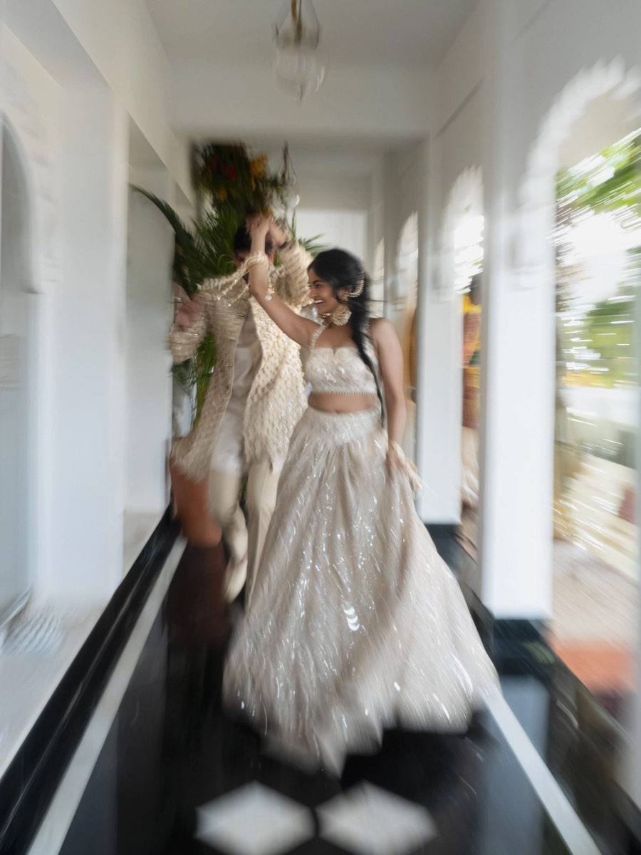 Two women in traditional Indian bridal attire dancing joyfully in a hallway.
