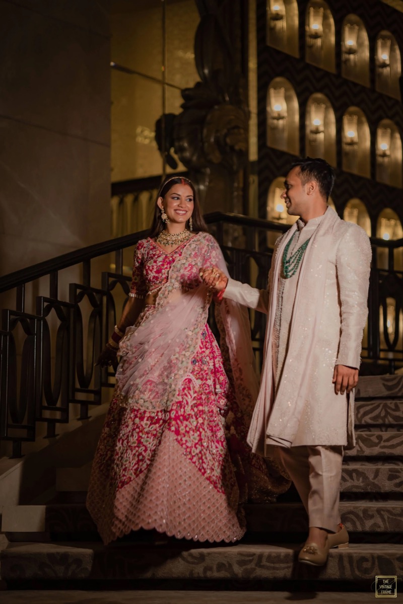 A bride and groom dressed in traditional Indian wedding attire, holding hands and smiling at each other on a staircase at an indoor venue with ornate lighting and decor.