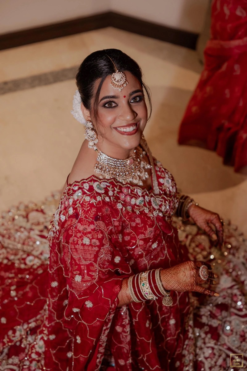 A smiling woman dressed in traditional red and gold Indian bridal attire, adorned with jewelry including earrings, necklace, and tikka, sitting on the floor with her hands decorated with henna.
