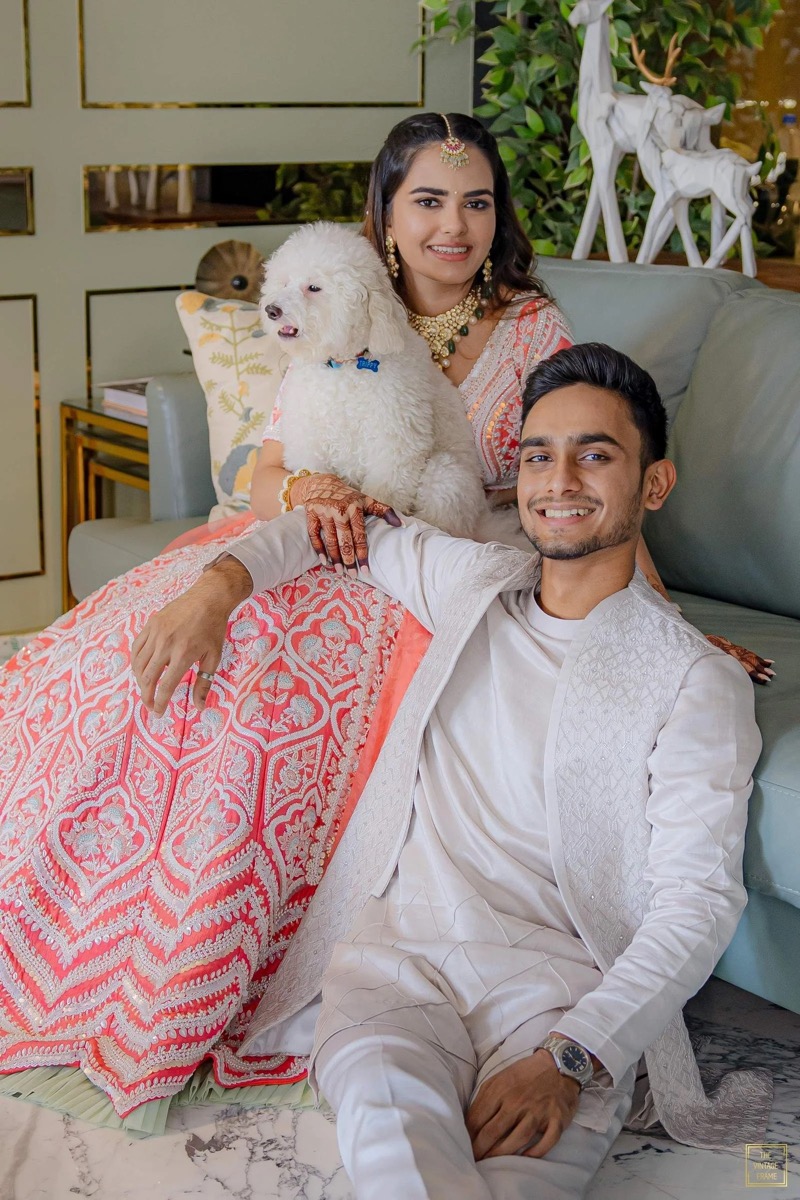 A smiling young man and woman in traditional Indian attire pose with a white poodle dog on a sofa in a decorated indoor setting.