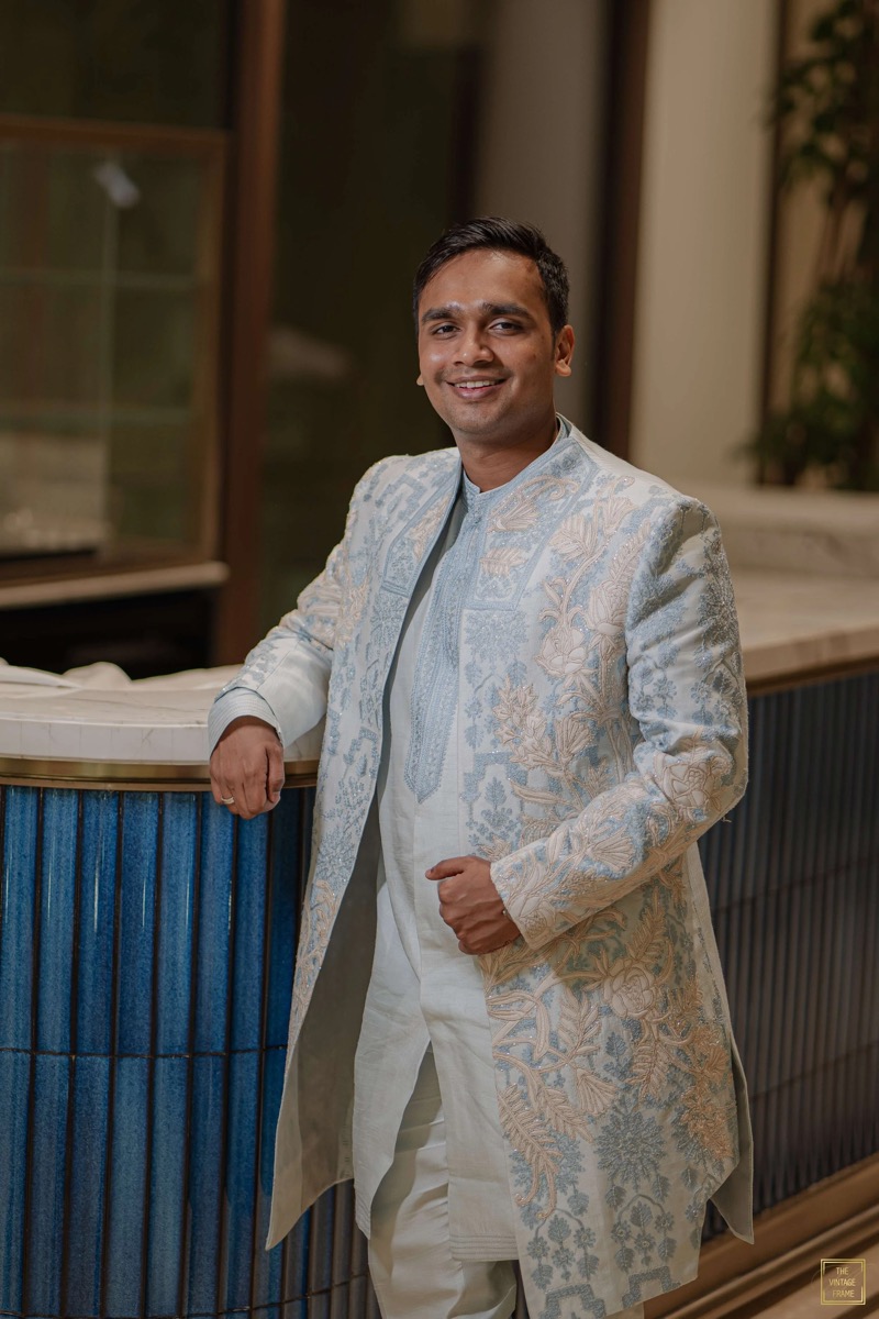 A man in a traditional Indian cream-colored sherwani with intricate floral embroidery, posing with a smile inside a room with wooden and glass elements.