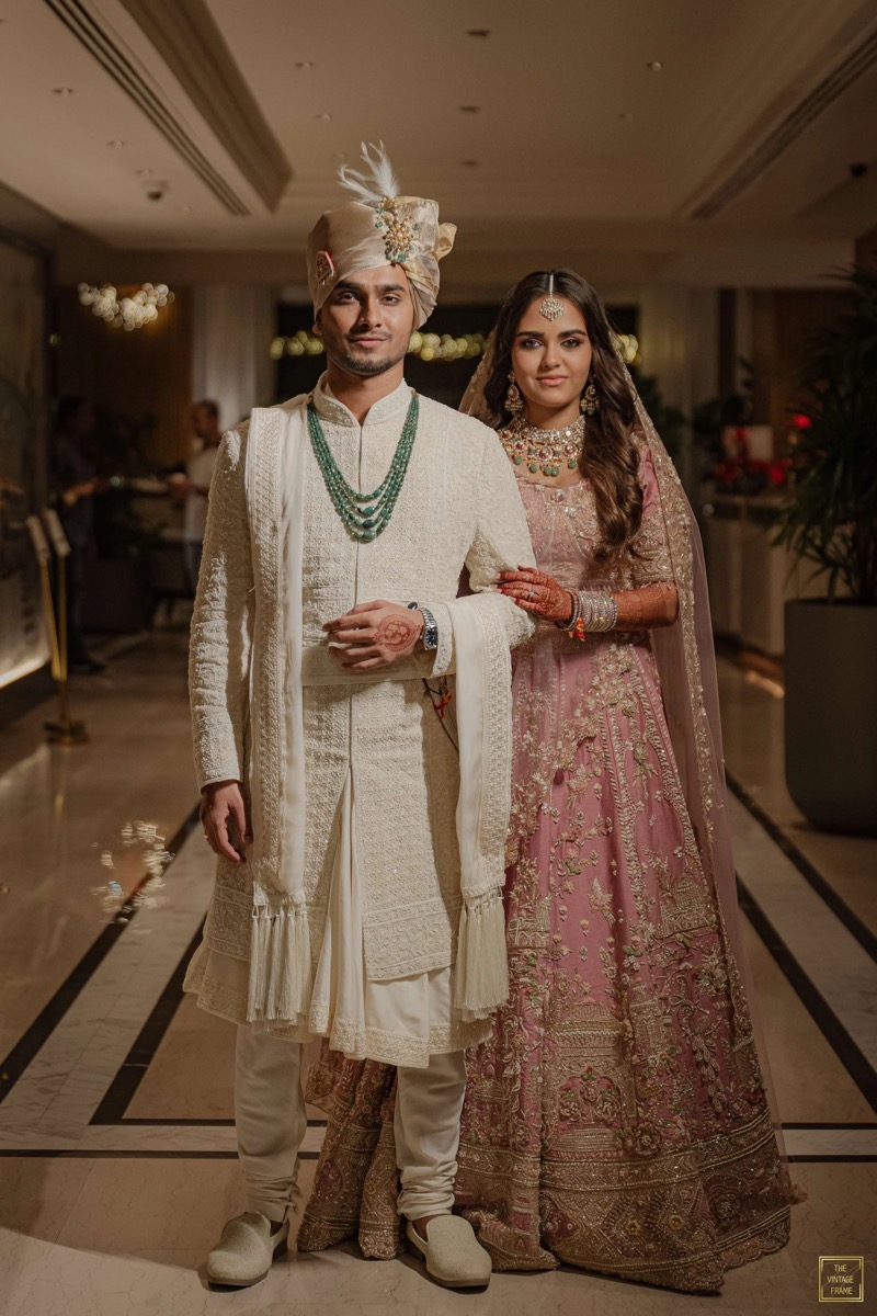 A couple dressed in traditional Indian wedding attire stands together in an elegant indoor setting, with the groom in cream-colored embroidery and a turban, and the bride in a pink and gold embroidered lehenga, both adorned with jewelry.