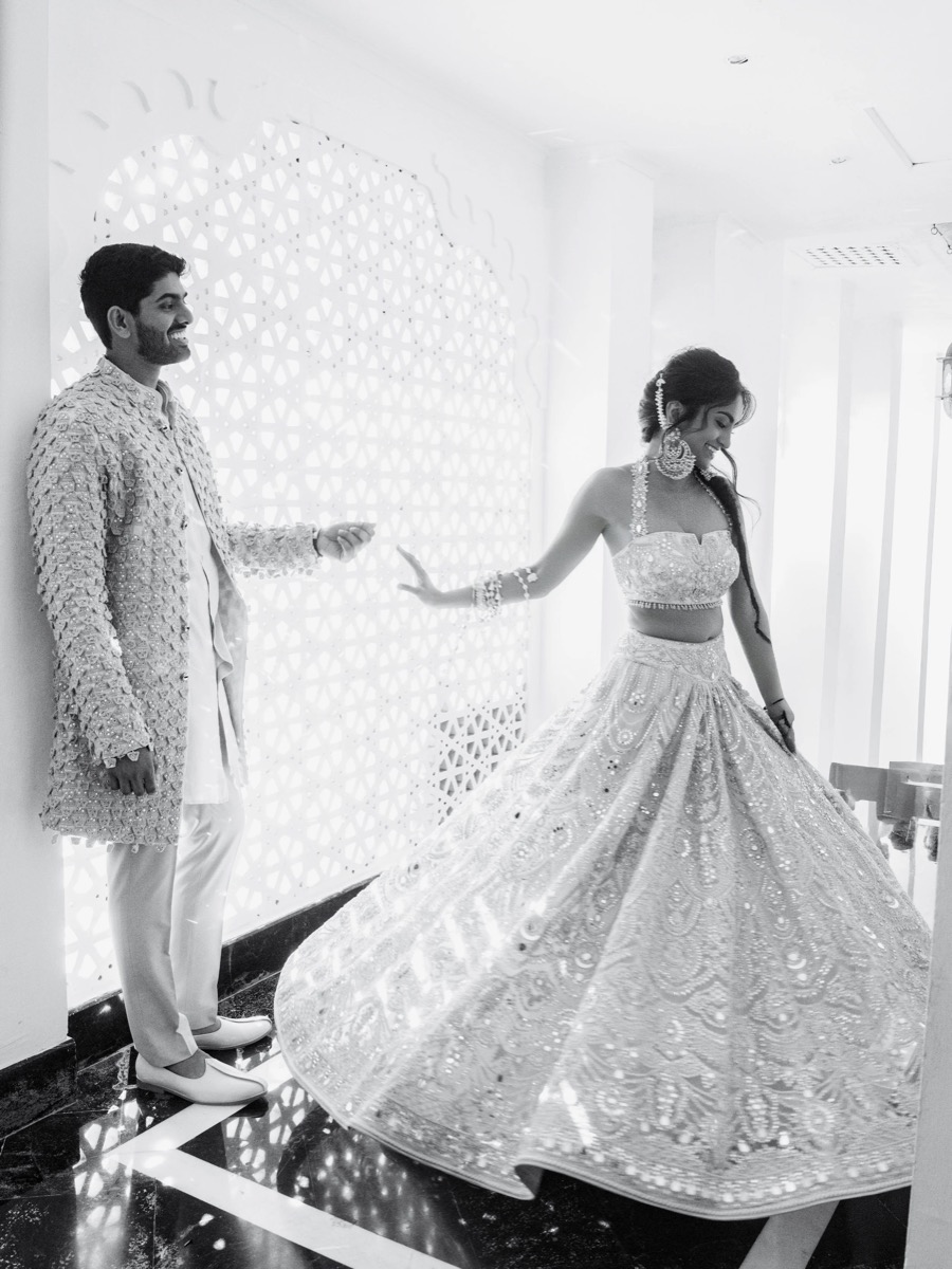 A man and woman dressed in traditional Indian wedding attire, smiling and dancing indoors.