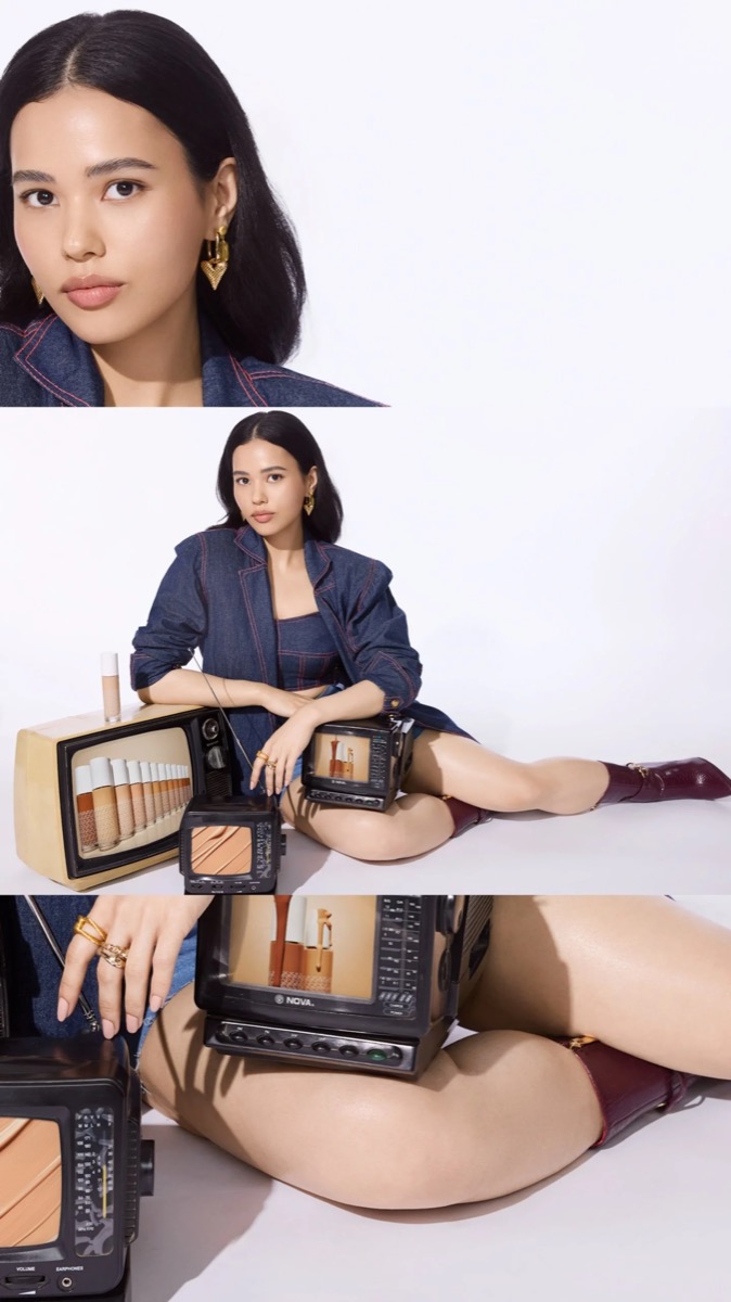A woman with long dark hair, wearing a denim jacket and gold earrings, sitting on the floor amidst vintage televisions and tubes of lipstick, with a white background.
