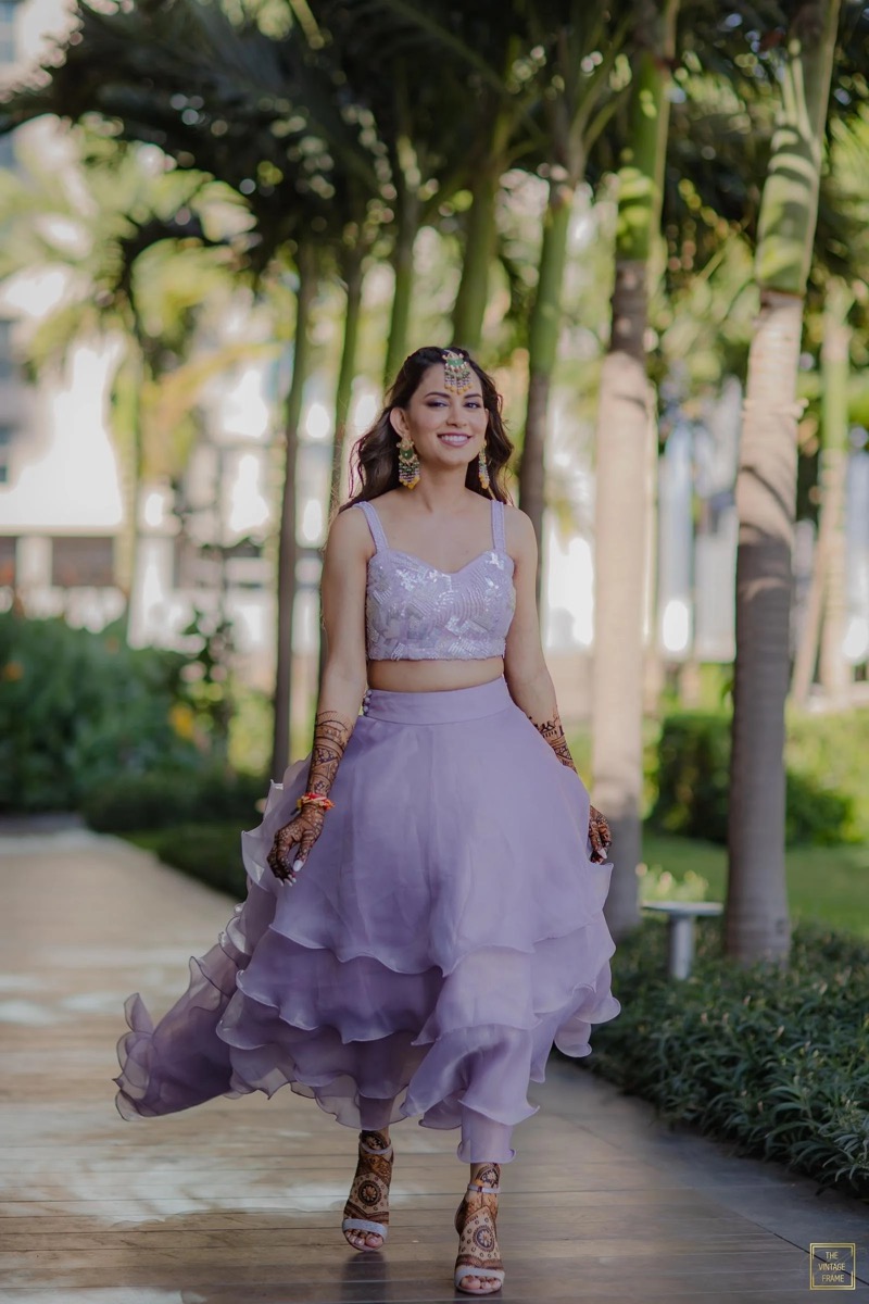 A woman dressed in lavender traditional Indian attire, with lotus and chicken embroidery, walking outdoors on a wooden pathway surrounded by palm trees, smiling.