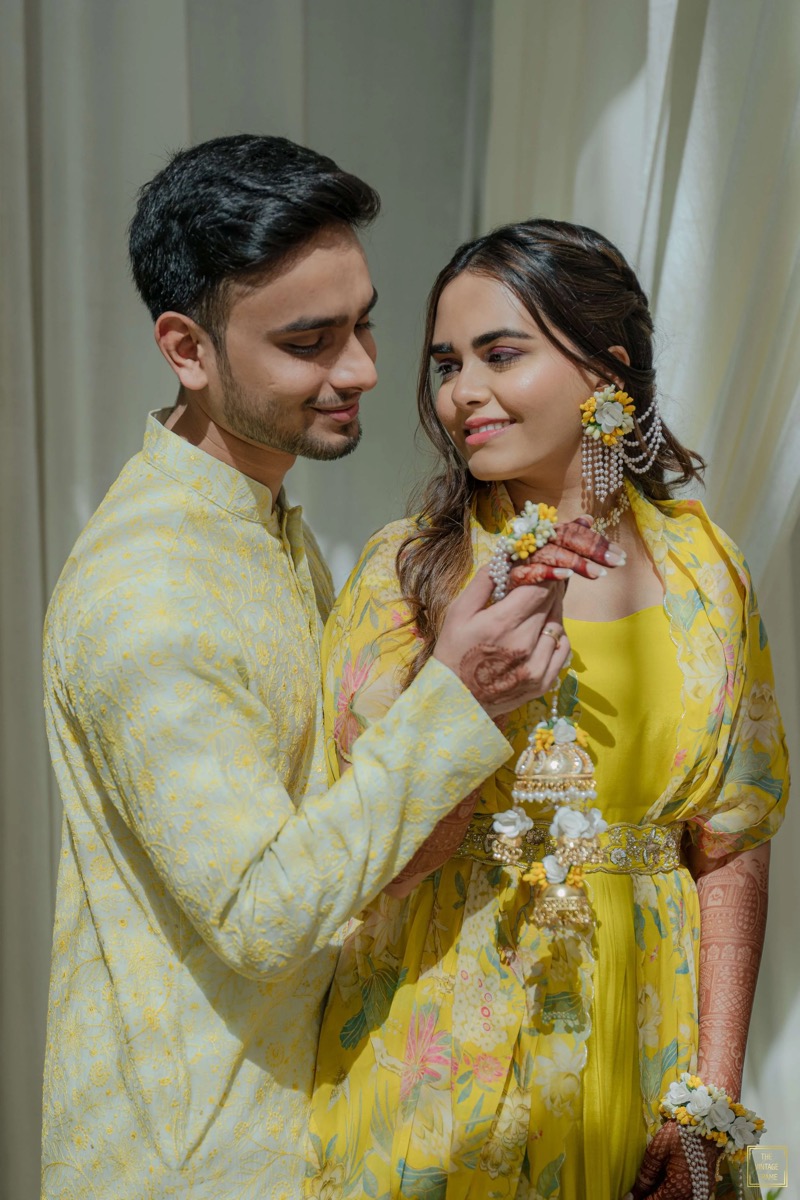 A couple dressed in yellow traditional attire, the woman with henna on her hand and elaborate jewelry, standing close together with the man holding a jewelry piece near the woman's face.
