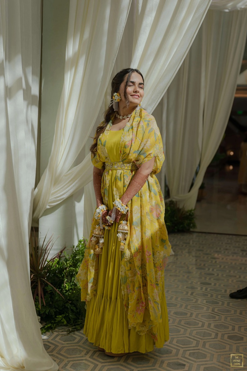 A woman dressed in a yellow floral dress with henna tattoos on her arms, standing in front of light-colored curtains, smiling with her eyes closed, holding jewelry in her hands.