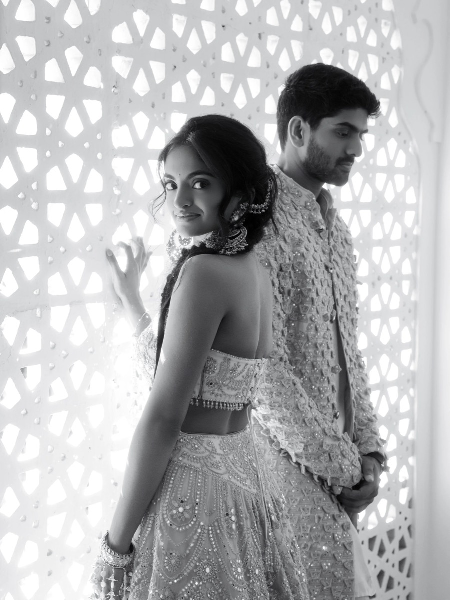 A woman and a man dressed in traditional Indian wedding attire, standing in front of a decorative screen. The woman is smiling and looking at the camera, while the man is looking downward with his hands clasped.