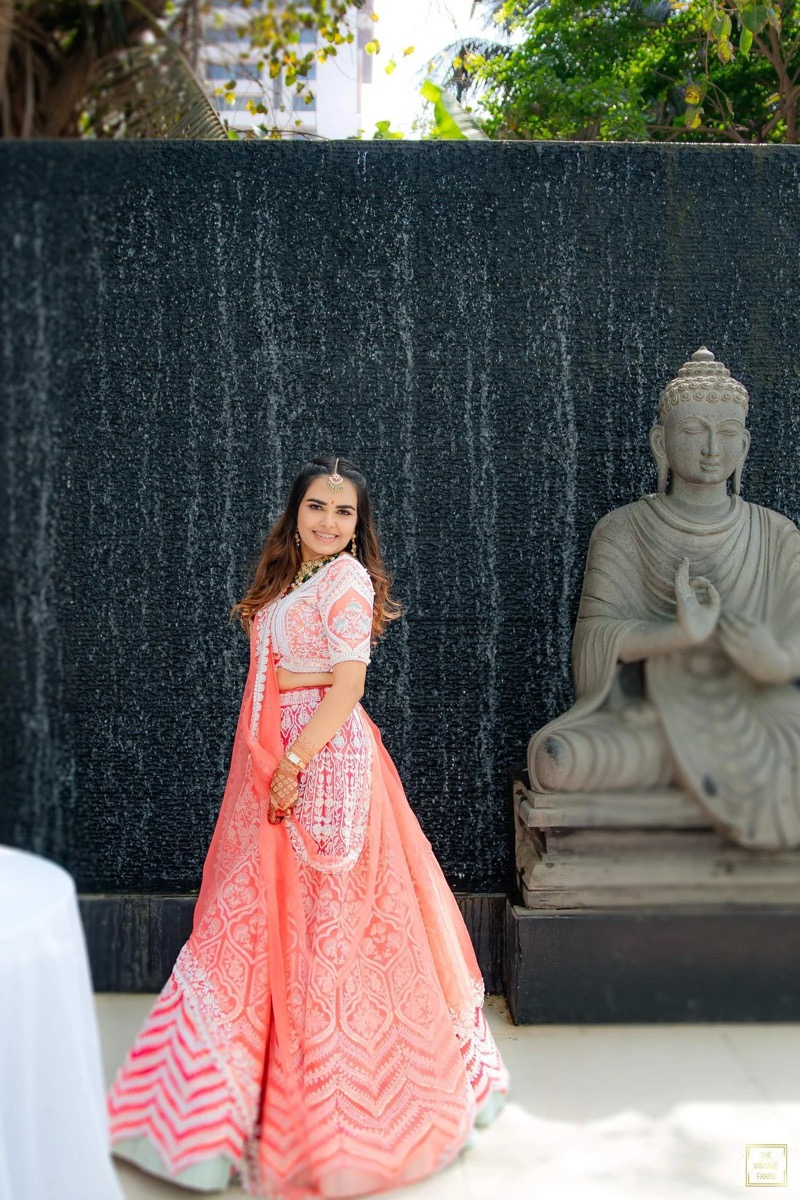 A smiling woman in a traditional pink and white Indian outfit with intricate embroidery, standing next to a Buddha statue at a decorative water fountain with a black textured wall and greenery in the background.