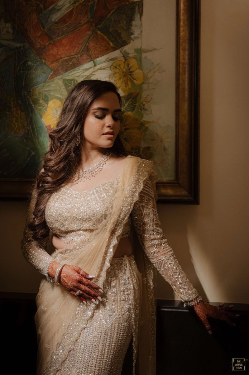 A woman dressed in a gold embroidered traditional Indian outfit, standing indoors against a beige wall with a framed colorful painting featuring flowers in the background.