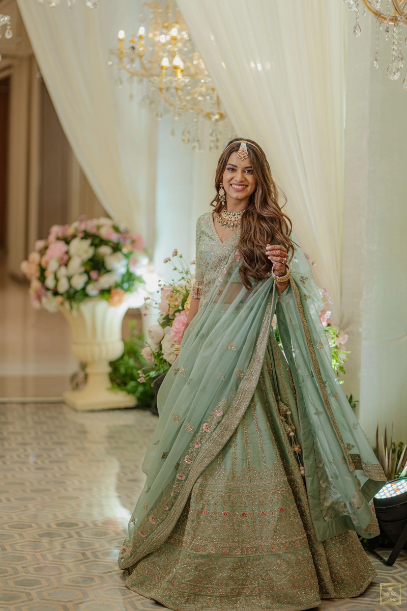 A woman dressed in a traditional, ornate green Indian outfit with gold embroidery, smiling at an event decorated with flowers and chandeliers.