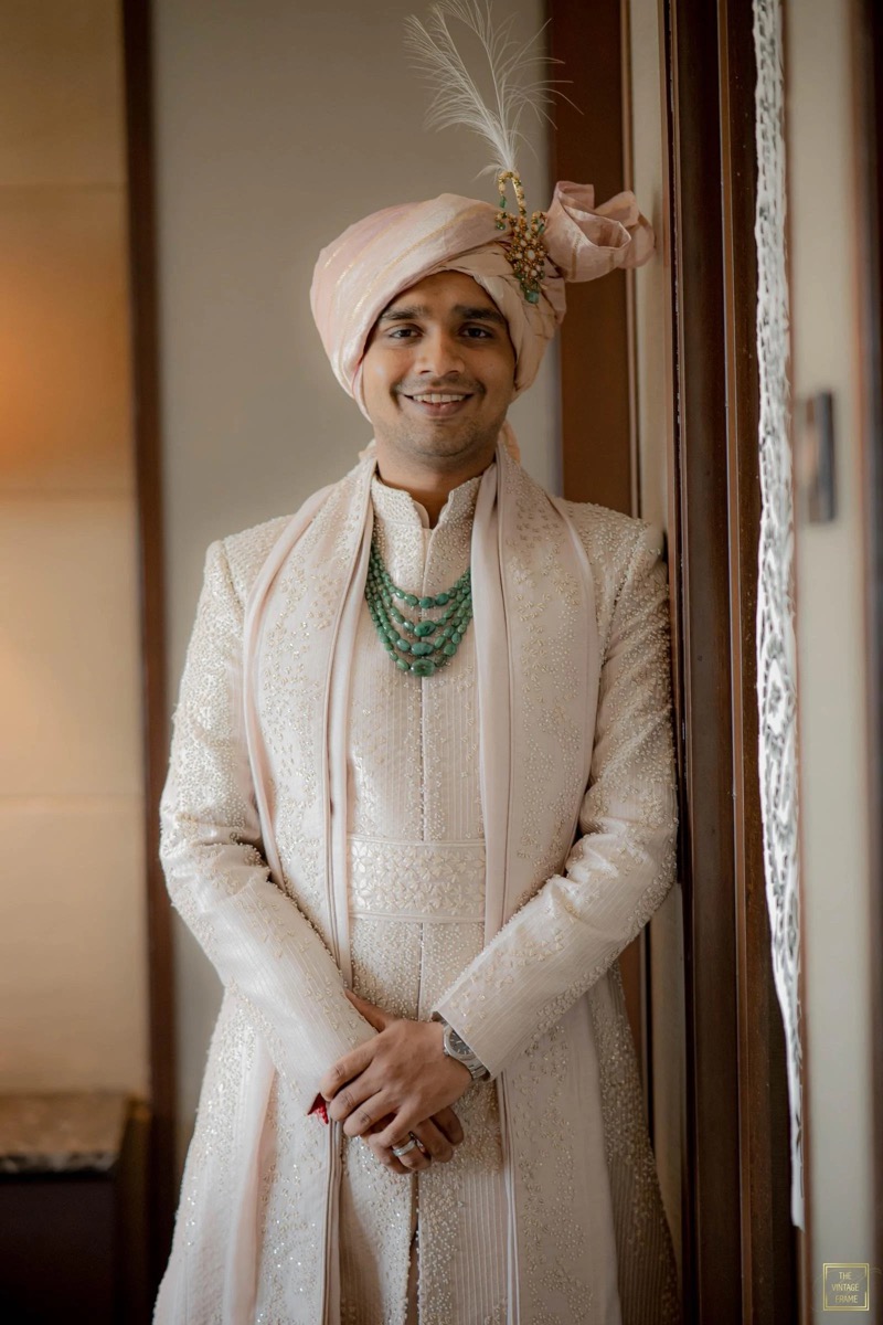 Man dressed in traditional Indian wedding attire, wearing a cream-colored sherwani with intricate embroidery, a matching turban with a feather and decorative embellishment, and layered turquoise necklaces, standing indoors next to a wooden wall.
