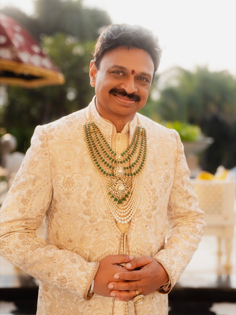 Man in traditional Indian wedding attire wearing multiple layered necklaces, standing outdoors with a warm smile.