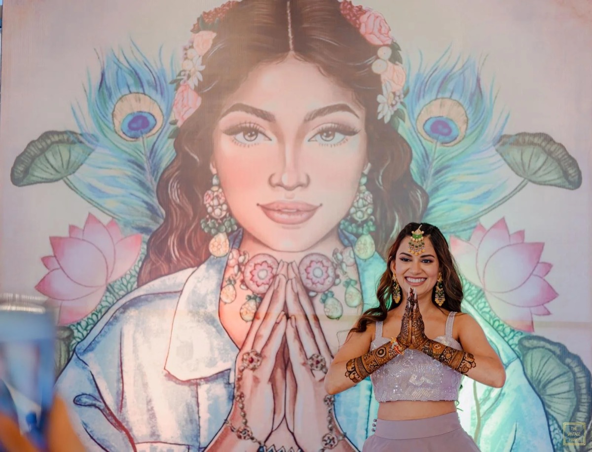 A woman dressed in traditional Indian attire standing in front of a large artistic mural of a woman with jewelry and a floral headdress. The woman has henna on her hands and is smiling with her hands in a prayer position.