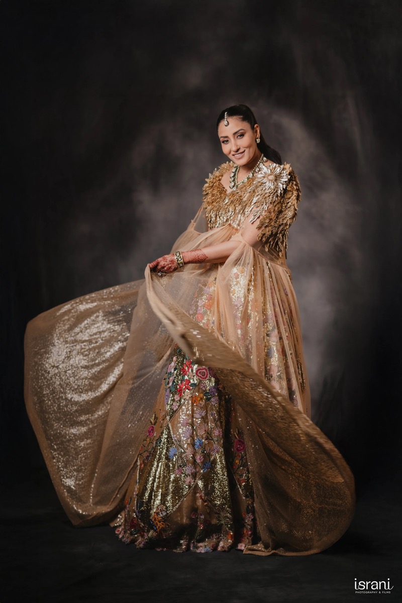 A woman dressed in an elaborate, embroidered gown with floral and gold embellishments poses against a dark backdrop.