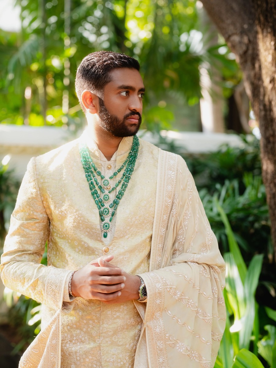 A man in traditional cream-colored Indian attire wearing multiple green beaded necklaces, standing outdoors with lush green foliage in the background.