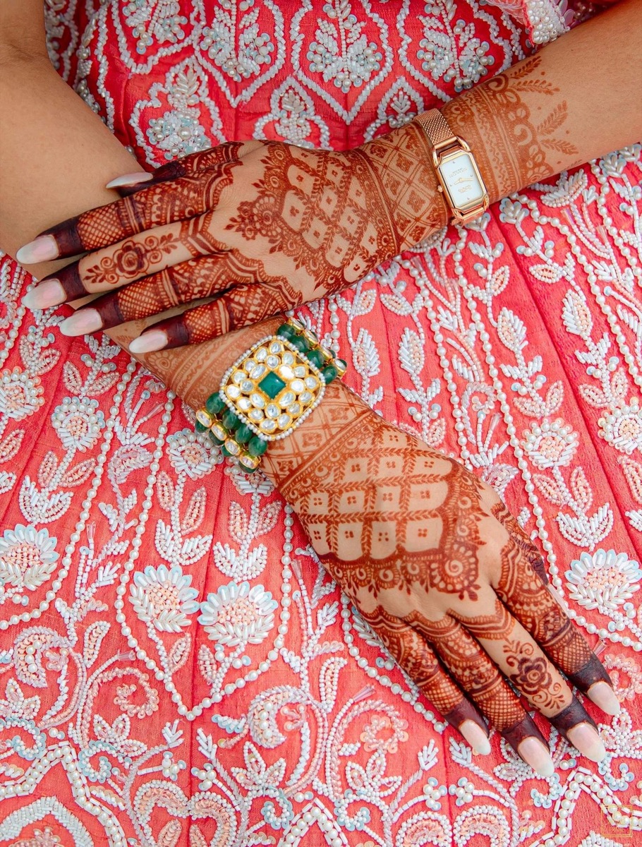 Close-up of hands and arms decorated with intricate henna designs resting on a red embroidered fabric. One hand has a bracelet and watch, and the fabric and henna feature traditional patterns and jewelry.