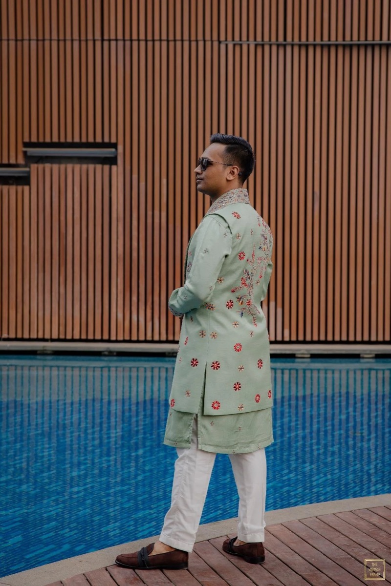 A man standing by a swimming pool, dressed in a light green embroidered kurta and white pants, wearing sunglasses, with a wooden wall in the background.