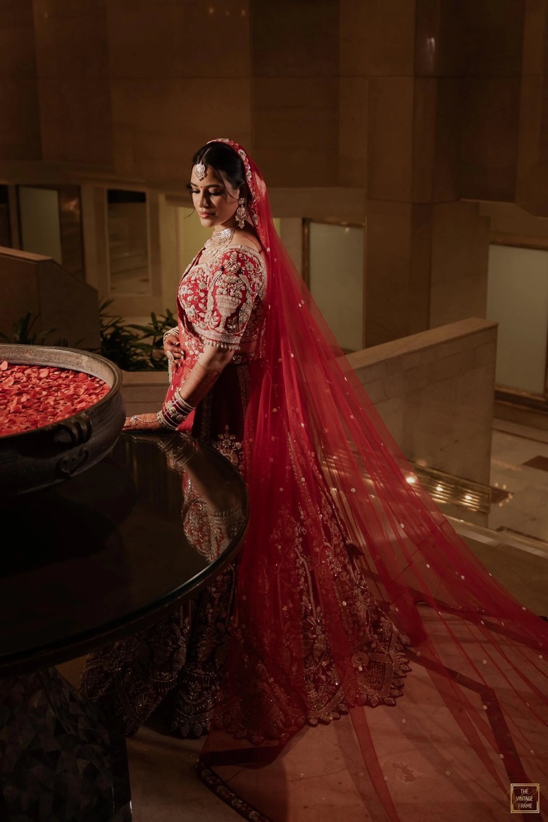 A woman dressed in a traditional red and gold bridal outfit with intricate embroidery, wearing jewelry and a veil, standing next to a large bowl of rose petals on a dark, polished table in an elegant indoor setting.