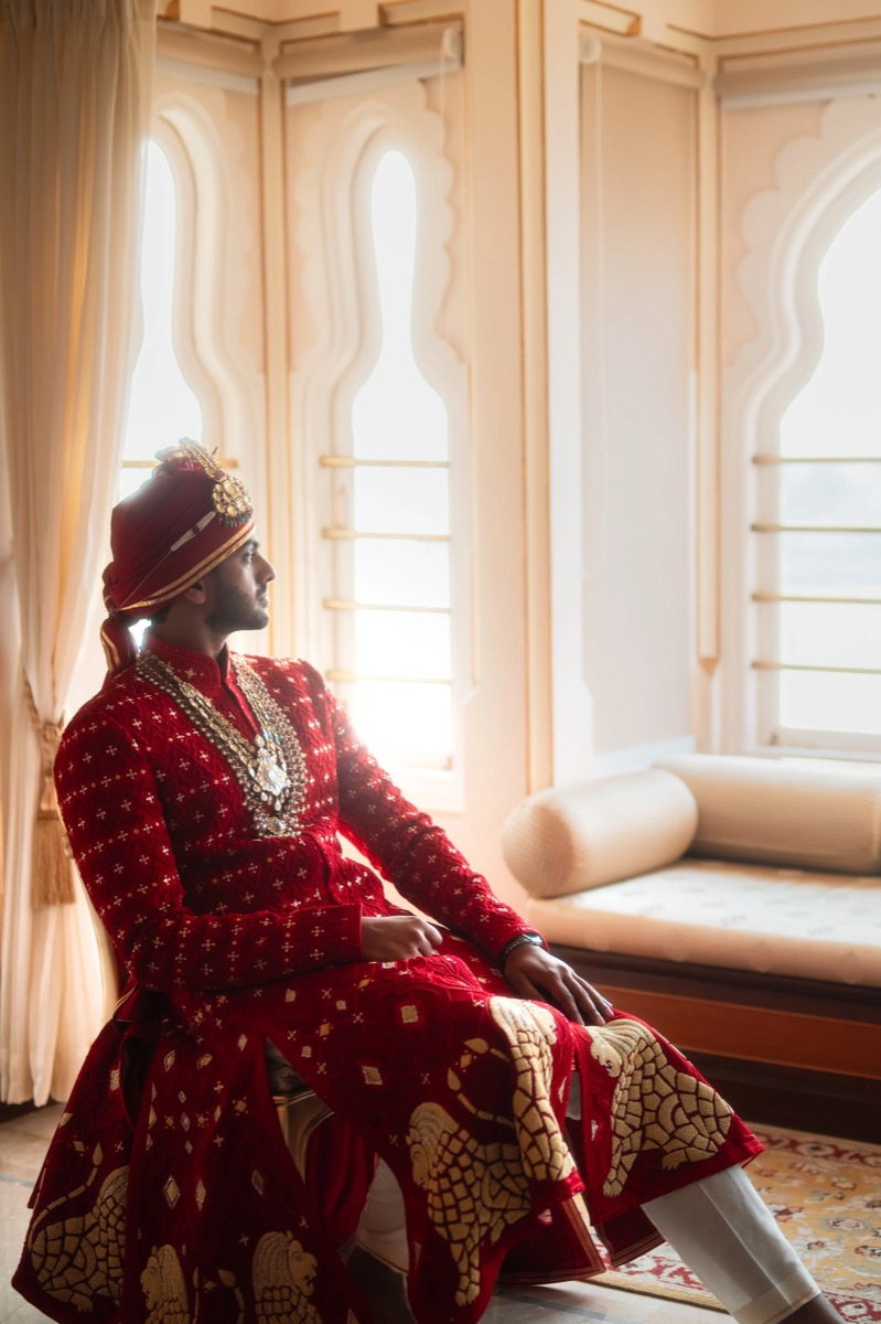 Man wearing traditional Indian wedding attire, including a red sherwani adorned with gold embroidery and a matching turban, sitting by the window.