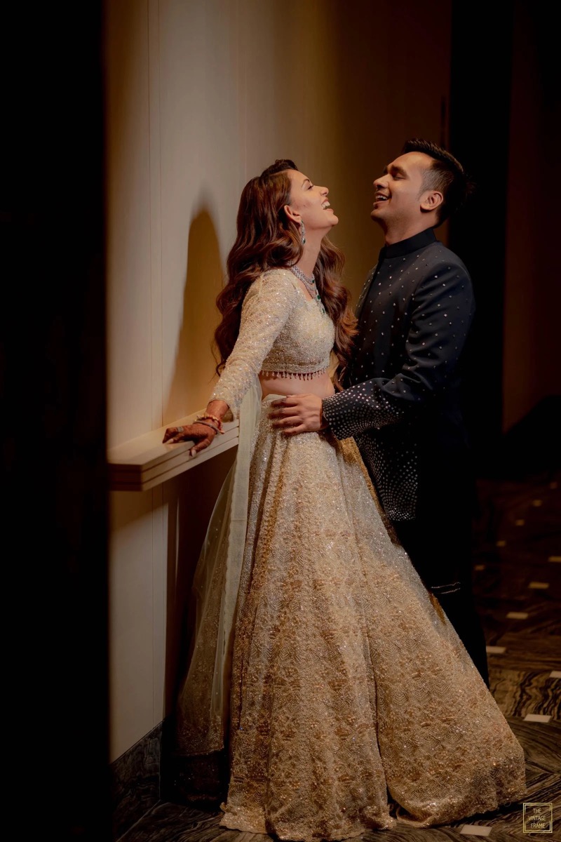 A couple dressed in traditional Indian wedding attire, laughing and looking at each other, standing indoors with a beige wall behind them.